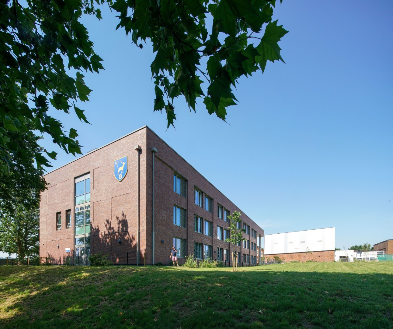 Modern three-story red brick school building with a blue crest on the facade, situated on a green lawn.